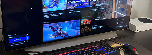 A computer desk with a monitor displaying the game menu of Overwatch 2, a keyboard, mouse, and a smartphone, set in a dimly lit room.